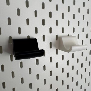 K&ouml;nnte beinhalten: Zwei kleine, minimalistische Regalhalterungen, eine schwarze und eine wei&szlig;e, sind an einer wei&szlig;en Lochwand befestigt. Die Halterungen haben ein einfaches, gebogenes Design, ideal zum Halten kleiner Gegenst&auml;nde. Die Lochwand hat gleichm&auml;&szlig;ig verteilte, ovale L&ouml;cher.
