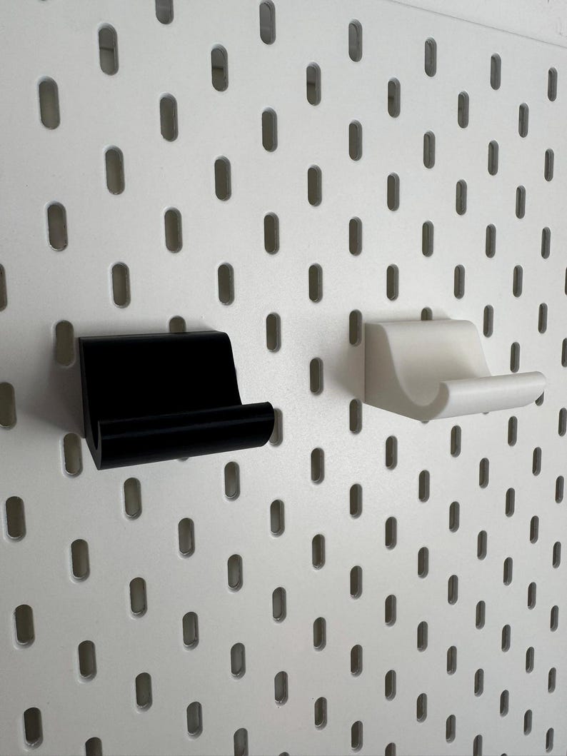 K&ouml;nnte beinhalten: Zwei kleine, minimalistische Regalhalterungen, eine schwarze und eine wei&szlig;e, sind an einer wei&szlig;en Lochwand befestigt. Die Halterungen haben ein einfaches, gebogenes Design, ideal zum Halten kleiner Gegenst&auml;nde. Die Lochwand hat gleichm&auml;&szlig;ig verteilte, ovale L&ouml;cher.