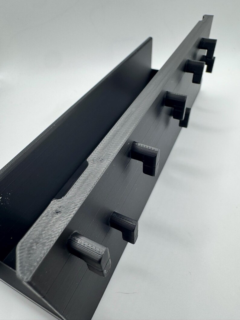 Peut inclure: Un organisateur rectangulaire noir avec un bord textur&eacute; et plusieurs crochets carr&eacute;s saillants. L'organisateur a un canal profond et est con&ccedil;u pour contenir des objets. L'objet est fait d'un mat&eacute;riau noir mat.