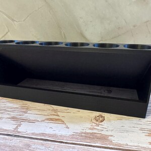 Puede incluir: Un organizador de madera negro con seis aberturas circulares en la parte superior y un compartimento rectangular debajo. El organizador est&aacute; en &aacute;ngulo, con un lado inclinado y una base plana. El art&iacute;culo est&aacute; sobre una superficie de madera blanca y marr&oacute;n desgastada.