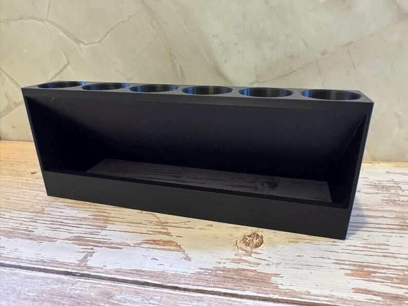 Puede incluir: Un organizador de madera negro con seis aberturas circulares en la parte superior y un compartimento rectangular debajo. El organizador est&aacute; en &aacute;ngulo, con un lado inclinado y una base plana. El art&iacute;culo est&aacute; sobre una superficie de madera blanca y marr&oacute;n desgastada.