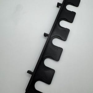 May include: A black, vertical rod holder with five curved slots for holding items. The holder has a rectangular shape with mounting tabs. The item is made of a textured material and is set against a white background.