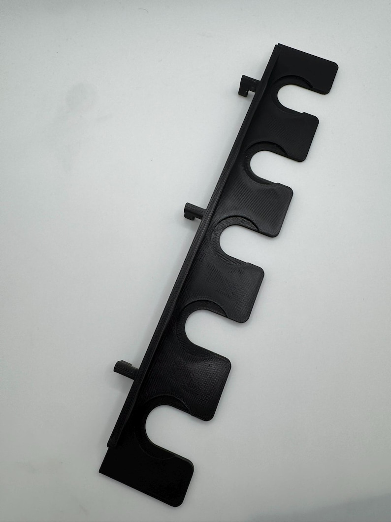 May include: A black, vertical rod holder with five curved slots for holding items. The holder has a rectangular shape with mounting tabs. The item is made of a textured material and is set against a white background.