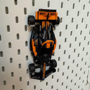 Op de afbeelding: Een zwarte en oranje Lego Formule 1 raceauto met het woord "Chrome" erop gedrukt. De auto hangt aan een wit prikbord.