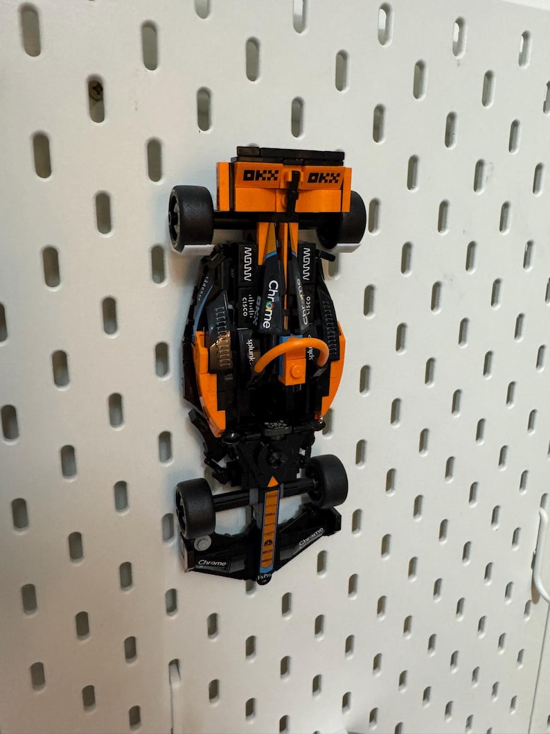 Op de afbeelding: Een zwarte en oranje Lego Formule 1 raceauto met het woord "Chrome" erop gedrukt. De auto hangt aan een wit prikbord.