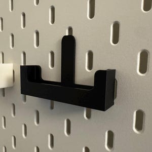 以下が含まれることがあります： 白黒のペグボードオーガナイザー。黒いオーガナイザーは、長方形のベースと垂直のサポートに焦点を当てています。左側には白いオーガナイザーがあります。どちらもペグボードの穴に合うように設計されています。