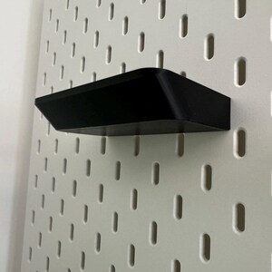 Puede incluir: Una estanter&iacute;a de pl&aacute;stico negro montada en una pared de tablero de clavijas blanco.