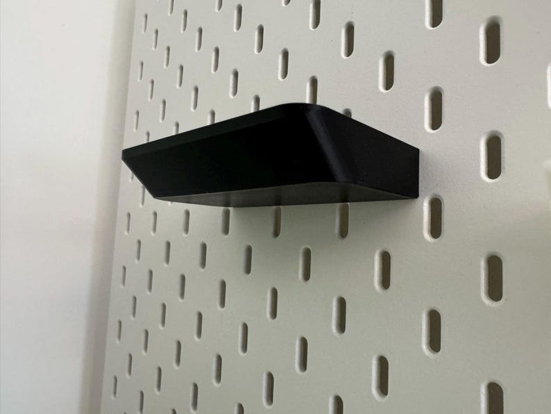 Puede incluir: Una estanter&iacute;a de pl&aacute;stico negro montada en una pared de tablero de clavijas blanco.
