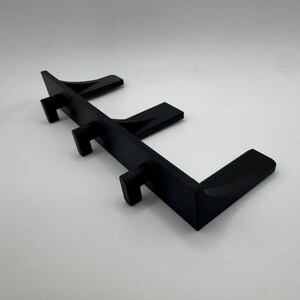 May include: A black, angled display stand with three vertical supports and a horizontal base. The stand is designed to hold items and is made of a solid, matte material. The design is modern and minimalist.