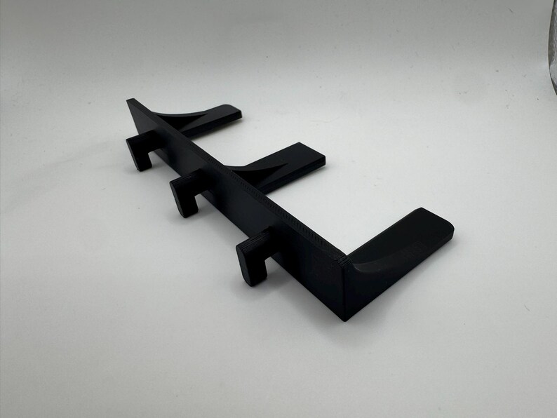 May include: A black, angled display stand with three vertical supports and a horizontal base. The stand is designed to hold items and is made of a solid, matte material. The design is modern and minimalist.