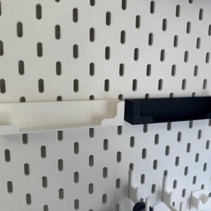 May include: White and black storage shelves designed for a pegboard system. The shelves are rectangular with a lip on the front to prevent items from falling out. The pegboard has evenly spaced holes.