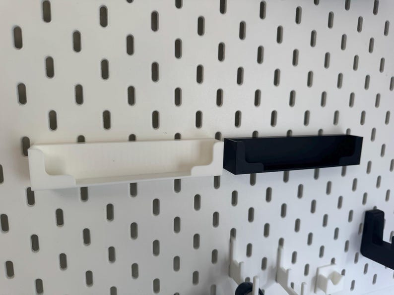 May include: White and black storage shelves designed for a pegboard system. The shelves are rectangular with a lip on the front to prevent items from falling out. The pegboard has evenly spaced holes.