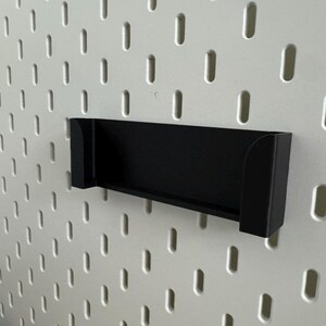 Pu&ograve; includere: Una mensola nera rettangolare progettata per una pannello forato. La mensola &egrave; montata su una pannello forato bianco con fori ovali distanziati uniformemente. Il design della mensola &egrave; semplice e moderno.