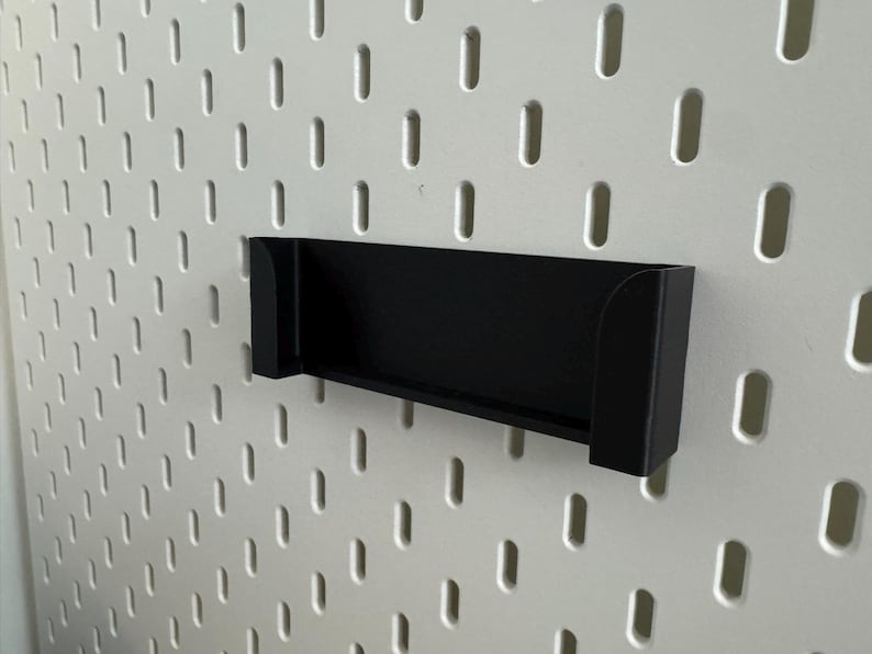 Pu&ograve; includere: Una mensola nera rettangolare progettata per una pannello forato. La mensola &egrave; montata su una pannello forato bianco con fori ovali distanziati uniformemente. Il design della mensola &egrave; semplice e moderno.