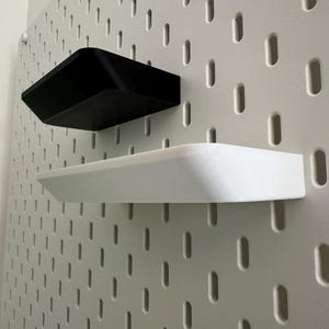 Puede incluir: Dos estantes montados en una pared de tablero de clavijas blanco. El estante superior es negro y el estante inferior es blanco.