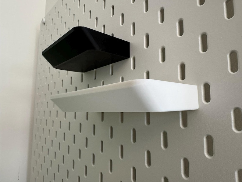 Puede incluir: Dos estantes montados en una pared de tablero de clavijas blanco. El estante superior es negro y el estante inferior es blanco.