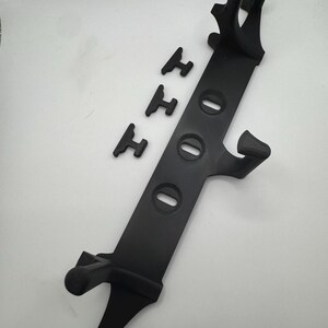May include: A black plastic stand with a long, rectangular body and three oval-shaped holes. The stand has two curved arms at the top and two legs at the bottom. Three small, black, H-shaped pieces are also included.