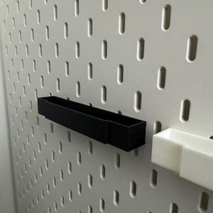 K&ouml;nnte beinhalten: Eine schwarze rechteckige Aufbewahrungsbox und eine wei&szlig;e rechteckige Aufbewahrungsbox sind an einer wei&szlig;en Lochwand befestigt. Die Lochwand hat gleichm&auml;&szlig;ig verteilte, vertikale Schlitze. Die schwarze Box ist l&auml;nger als die wei&szlig;e Box.