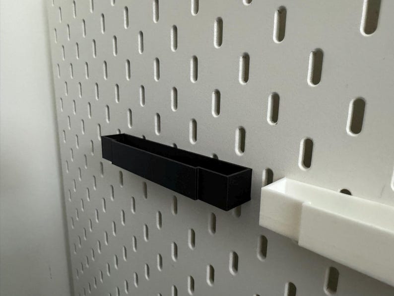K&ouml;nnte beinhalten: Eine schwarze rechteckige Aufbewahrungsbox und eine wei&szlig;e rechteckige Aufbewahrungsbox sind an einer wei&szlig;en Lochwand befestigt. Die Lochwand hat gleichm&auml;&szlig;ig verteilte, vertikale Schlitze. Die schwarze Box ist l&auml;nger als die wei&szlig;e Box.