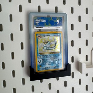 Op de afbeelding: Een beoordeelde Pok&eacute;mon-kaart met Vaporeon uit de Jungle-set van 1999. De kaart zit in een doorzichtige houder en wordt tentoongesteld op een zwarte standaard. De kaart is beoordeeld met VG 4.