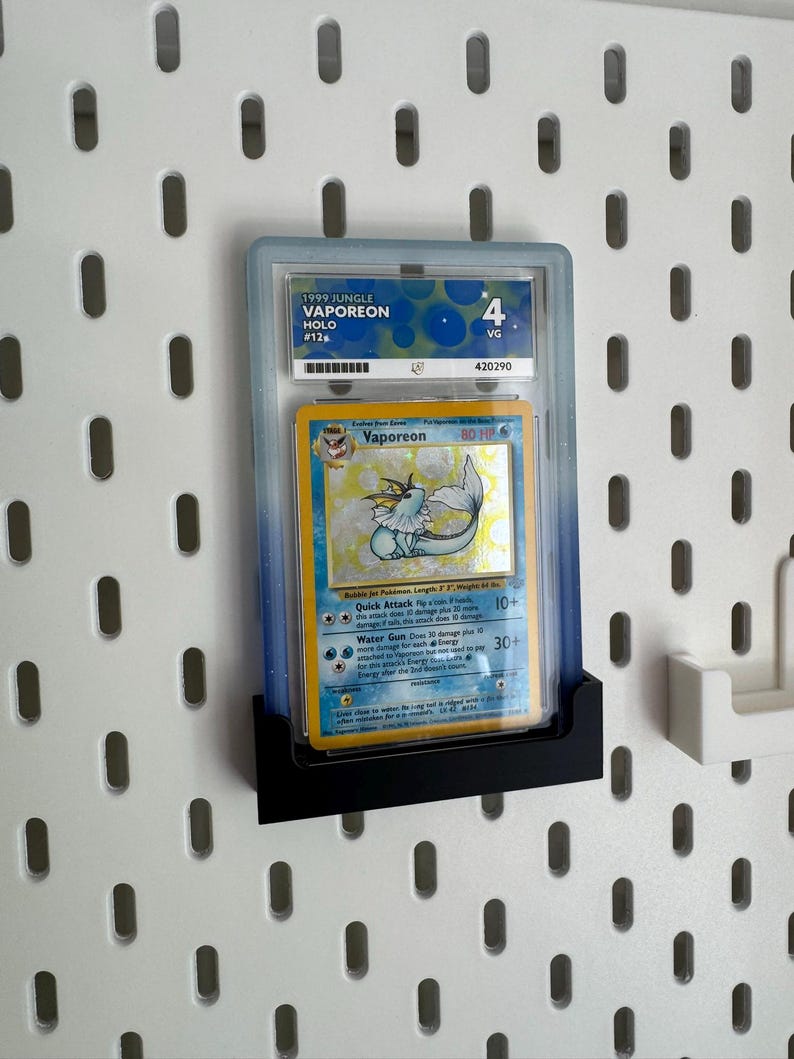 Op de afbeelding: Een beoordeelde Pok&eacute;mon-kaart met Vaporeon uit de Jungle-set van 1999. De kaart zit in een doorzichtige houder en wordt tentoongesteld op een zwarte standaard. De kaart is beoordeeld met VG 4.