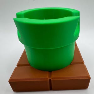 May include: A green pipe, resembling a video game element, rests on a brown brick-like base. The pipe has a rounded top and a cylindrical form. The base is made of four rectangular blocks. The objects are made of plastic.