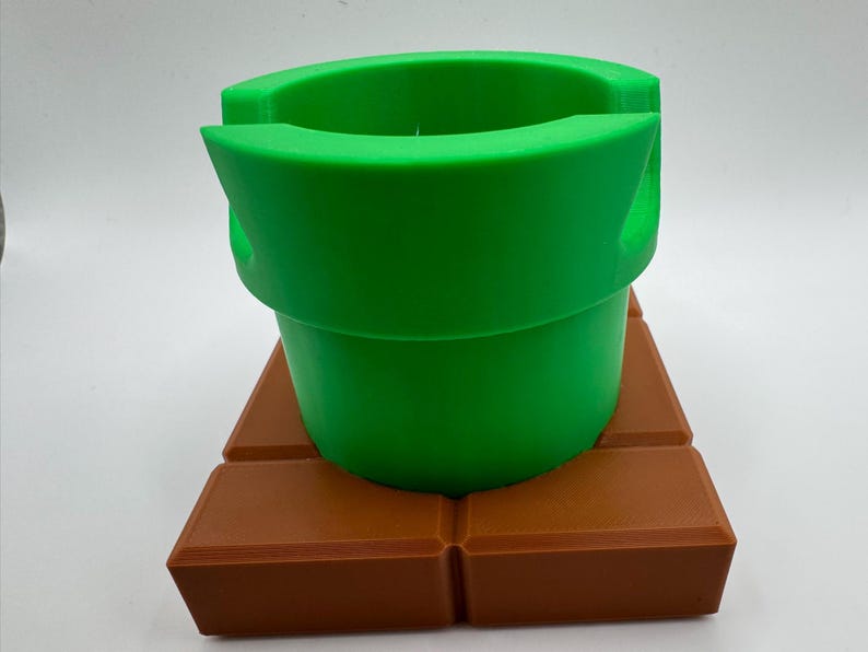 May include: A green pipe, resembling a video game element, rests on a brown brick-like base. The pipe has a rounded top and a cylindrical form. The base is made of four rectangular blocks. The objects are made of plastic.