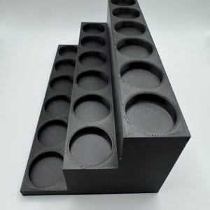 Puede incluir: Un soporte negro escalonado para botes de pintura con tres niveles, cada uno con m&uacute;ltiples aberturas circulares. El soporte est&aacute; hecho de un material negro mate s&oacute;lido y est&aacute; dise&ntilde;ado para contener botellas o recipientes peque&ntilde;os.