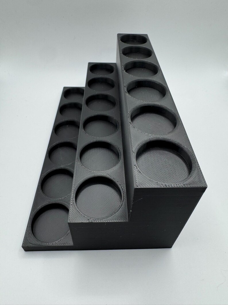 Puede incluir: Un soporte negro escalonado para botes de pintura con tres niveles, cada uno con m&uacute;ltiples aberturas circulares. El soporte est&aacute; hecho de un material negro mate s&oacute;lido y est&aacute; dise&ntilde;ado para contener botellas o recipientes peque&ntilde;os.
