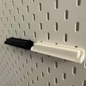 Pode incluir: Duas prateleiras de painel perfurado, uma preta e outra branca, s&atilde;o exibidas contra um painel perfurado branco. As prateleiras t&ecirc;m formato retangular com uma borda frontal curva. O painel perfurado tem orif&iacute;cios oblongos espa&ccedil;ados uniformemente.
