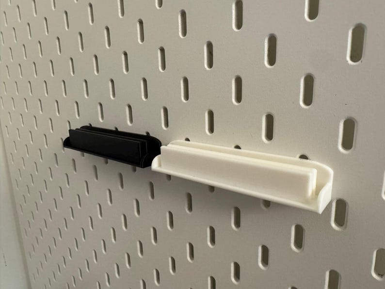 Pode incluir: Duas prateleiras de painel perfurado, uma preta e outra branca, s&atilde;o exibidas contra um painel perfurado branco. As prateleiras t&ecirc;m formato retangular com uma borda frontal curva. O painel perfurado tem orif&iacute;cios oblongos espa&ccedil;ados uniformemente.