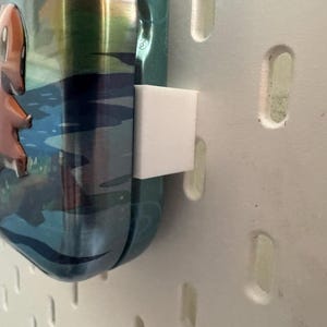 K&ouml;nnte beinhalten: Eine teal- und silberfarbene Wasserflasche mit einem Cartoon-Charakter von Charmander. Die Flasche wird von einem wei&szlig;en, quadratischen Halter gehalten, der an einer wei&szlig;en Lochplatte befestigt ist. Der Charmander ist orange und der Hintergrund hat eine blaue Wasserszene.
