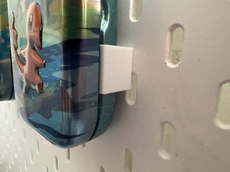 K&ouml;nnte beinhalten: Eine teal- und silberfarbene Wasserflasche mit einem Cartoon-Charakter von Charmander. Die Flasche wird von einem wei&szlig;en, quadratischen Halter gehalten, der an einer wei&szlig;en Lochplatte befestigt ist. Der Charmander ist orange und der Hintergrund hat eine blaue Wasserszene.