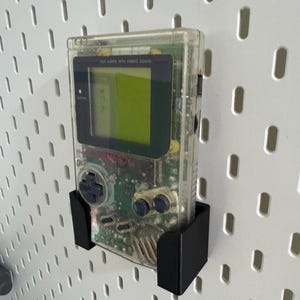 Op de afbeelding: Een transparante Game Boy Color handheld-videogamesysteem met een zwart en groen circuitbord dat zichtbaar is in het interieur. De console is gemonteerd op een zwarte pegboardhaak.