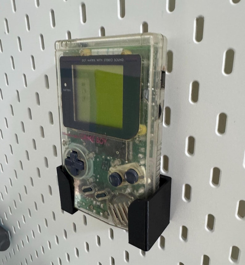 Op de afbeelding: Een transparante Game Boy Color handheld-videogamesysteem met een zwart en groen circuitbord dat zichtbaar is in het interieur. De console is gemonteerd op een zwarte pegboardhaak.