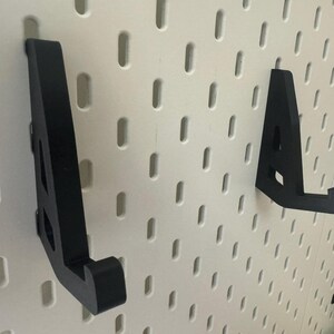 May include: Two black plastic wall mounted hooks for hanging items on a white pegboard.