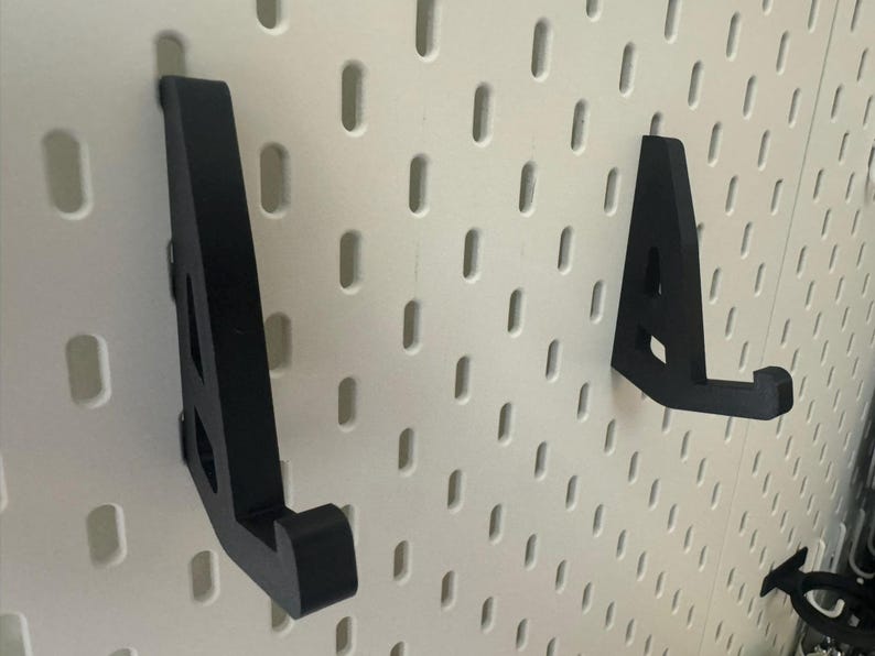 May include: Two black plastic wall mounted hooks for hanging items on a white pegboard.