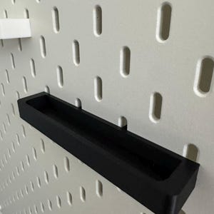 May include: A black rectangular shelf, designed to attach to a pegboard. The shelf is made of a solid, matte black material and has a recessed area for holding items. The pegboard is white with evenly spaced, oval-shaped holes.