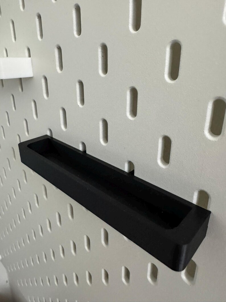 May include: A black rectangular shelf, designed to attach to a pegboard. The shelf is made of a solid, matte black material and has a recessed area for holding items. The pegboard is white with evenly spaced, oval-shaped holes.