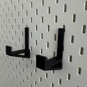 May include: Two black metal pegboard hooks are mounted on a white pegboard. The hooks are L-shaped and designed to hold items. The pegboard has evenly spaced holes for attaching the hooks.