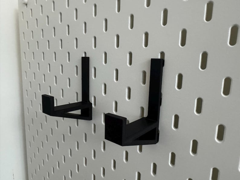 May include: Two black metal pegboard hooks are mounted on a white pegboard. The hooks are L-shaped and designed to hold items. The pegboard has evenly spaced holes for attaching the hooks.