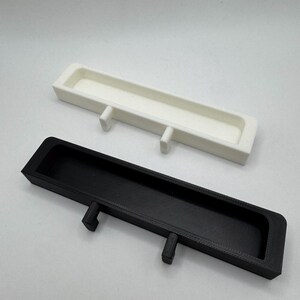May include: Two rectangular trays, one white and one black, are displayed. Each tray has a long, narrow trough and two small, rectangular legs. The trays are likely designed for organizing small items or as decorative elements.