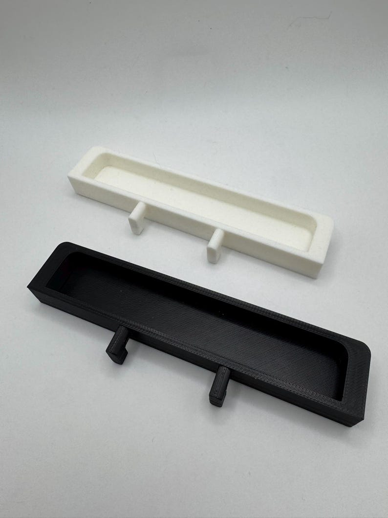 May include: Two rectangular trays, one white and one black, are displayed. Each tray has a long, narrow trough and two small, rectangular legs. The trays are likely designed for organizing small items or as decorative elements.
