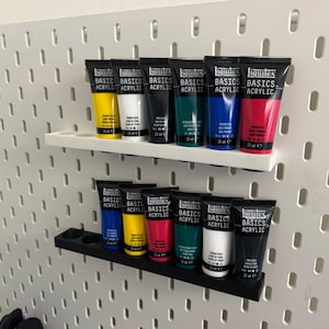 Può includere: Due mensole orizzontali montate su un pannello forato bianco, ciascuna contenente sei tubi di vernice acrilica Liquitex Basics. La mensola superiore è bianca e quella inferiore è nera, entrambe con fori circolari per i tubi.