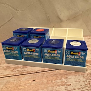 Peut inclure: Un organisateur en plastique blanc contenant six petits pots de peinture Revell Aqua Color bleus. Chaque pot est &eacute;tiquet&eacute; avec le nom de la marque, le type de peinture et un volume de 18 ml. Les pots sont dispos&eacute;s sur une surface en bois.