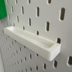 May include: A white, rectangular shelf attached to a pegboard. The shelf is made of a solid, opaque material and has a shallow, open top. The pegboard is white with evenly spaced, oblong holes. A green object is visible on the left.