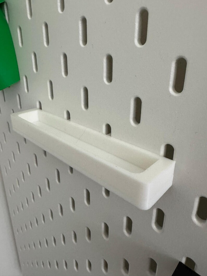 May include: A white, rectangular shelf attached to a pegboard. The shelf is made of a solid, opaque material and has a shallow, open top. The pegboard is white with evenly spaced, oblong holes. A green object is visible on the left.