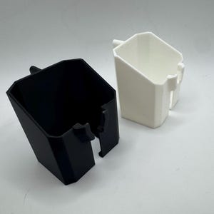 May include: Two geometric, angular containers. One is black, the other is white. Both have a rectangular shape with a handle-like protrusion on the side. The containers are likely for storage or organization.