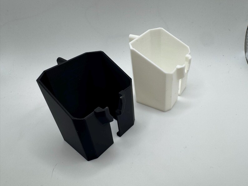 May include: Two geometric, angular containers. One is black, the other is white. Both have a rectangular shape with a handle-like protrusion on the side. The containers are likely for storage or organization.