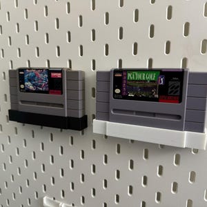 K&ouml;nnte beinhalten: Zwei graue Super Nintendo-Spielmodule. Eines hat einen schwarzen Sockel, das andere einen wei&szlig;en Sockel. Die Spielmodule sind an einer wei&szlig;en Lochwand befestigt. Ein Modul tr&auml;gt den Text "PGA TOUR GOLF".